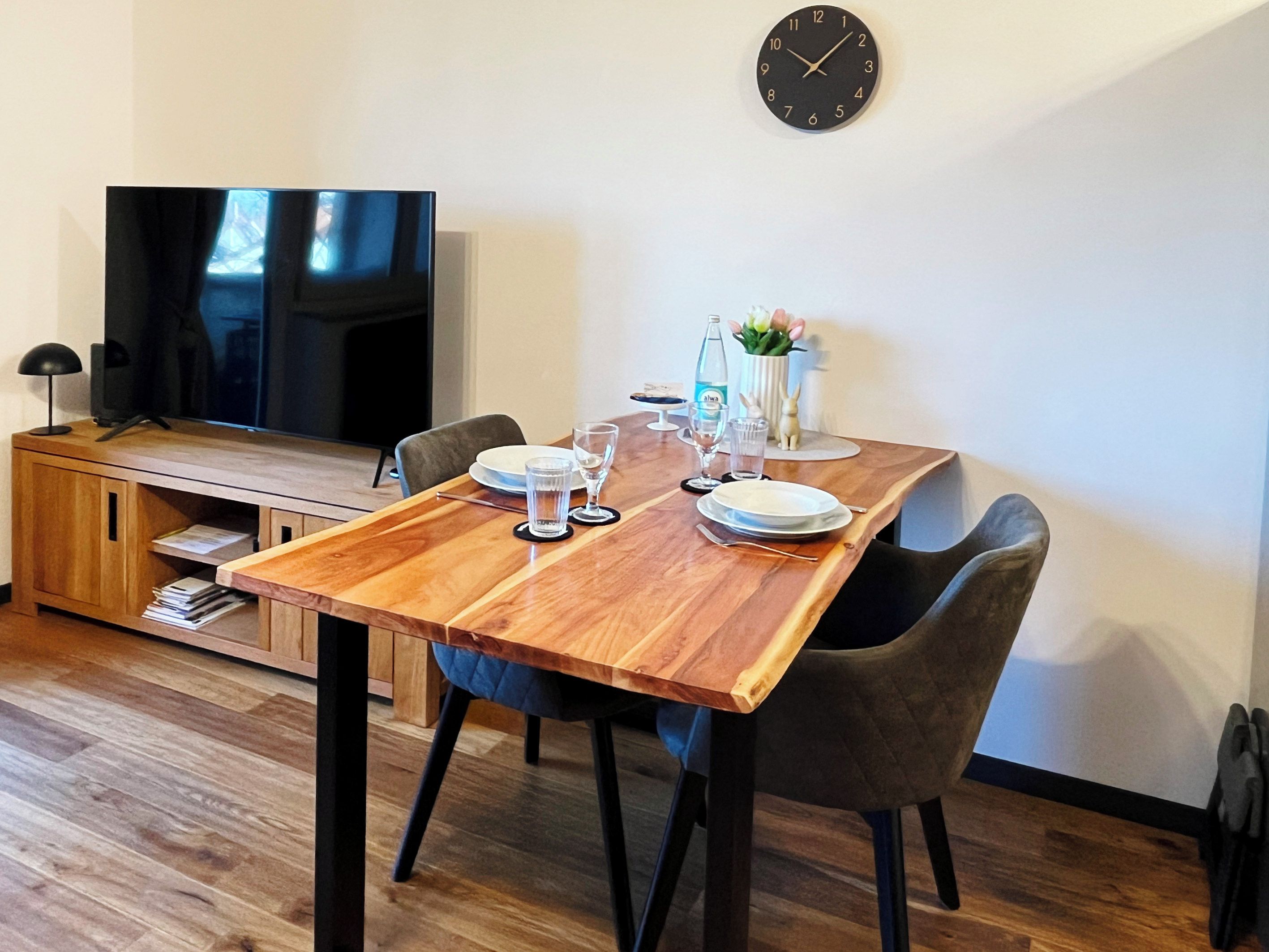 Ein Essbereich mit einem Holztisch, gedeckt für zwei Personen, einem Fernseher auf einem Schrank und einer Uhr an der Wand.