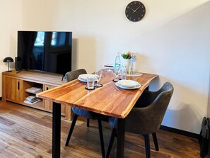 A dining area with a wooden table set for two, a television on a stand, and a clock on the wall.