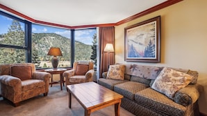 Un salon chaleureux avec une grande fenêtre offrant une vue imprenable sur les montagnes et les arbres. On y trouve une table basse en bois, deux fauteuils et un canapé agrémenté de coussins à motifs.