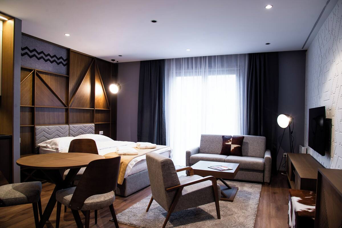 Una habitación de hotel moderna con cama, sofá, mesa de comedor, sillas y televisor.