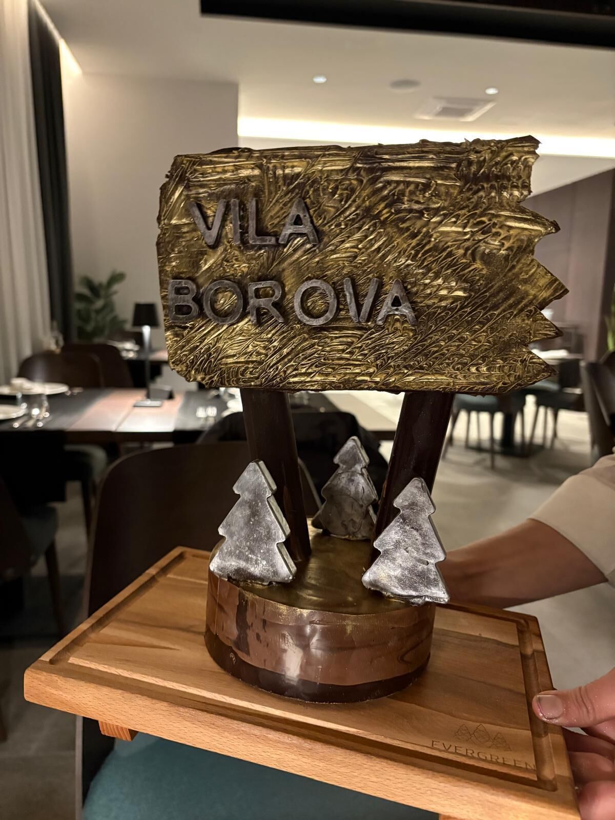 Een houten bord met de tekst 'VLA BOROVA' erop, ondersteund door drie zilveren kerstboomornamenten.