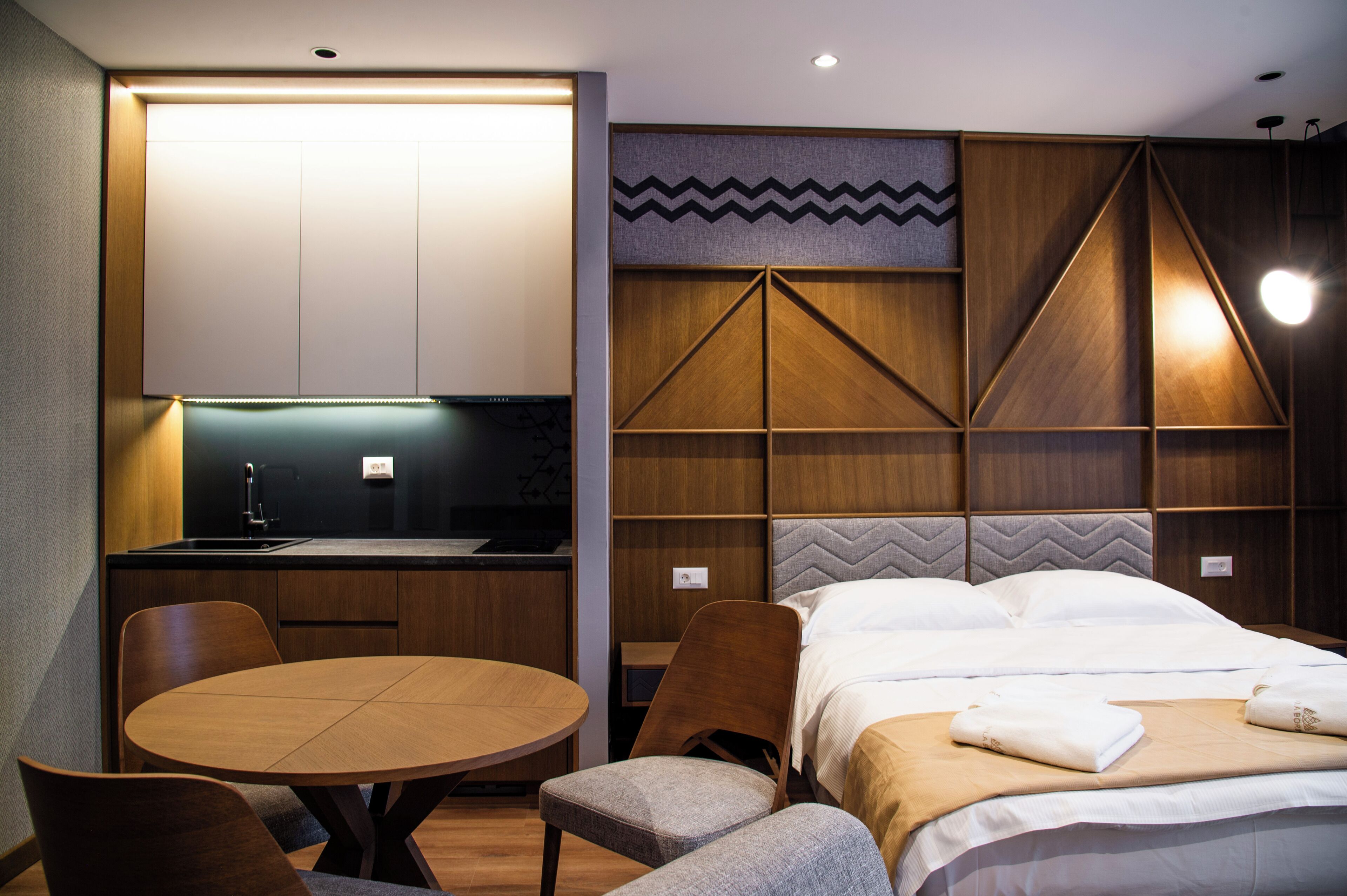 A modern hotel room with a kitchenette, a round dining table, and a bed with neatly made linens.