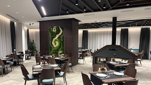 Un comedor moderno con varias mesas y sillas, una pared decorativa con estampado verde y blanco, y un techo con vigas a la vista e iluminación empotrada.