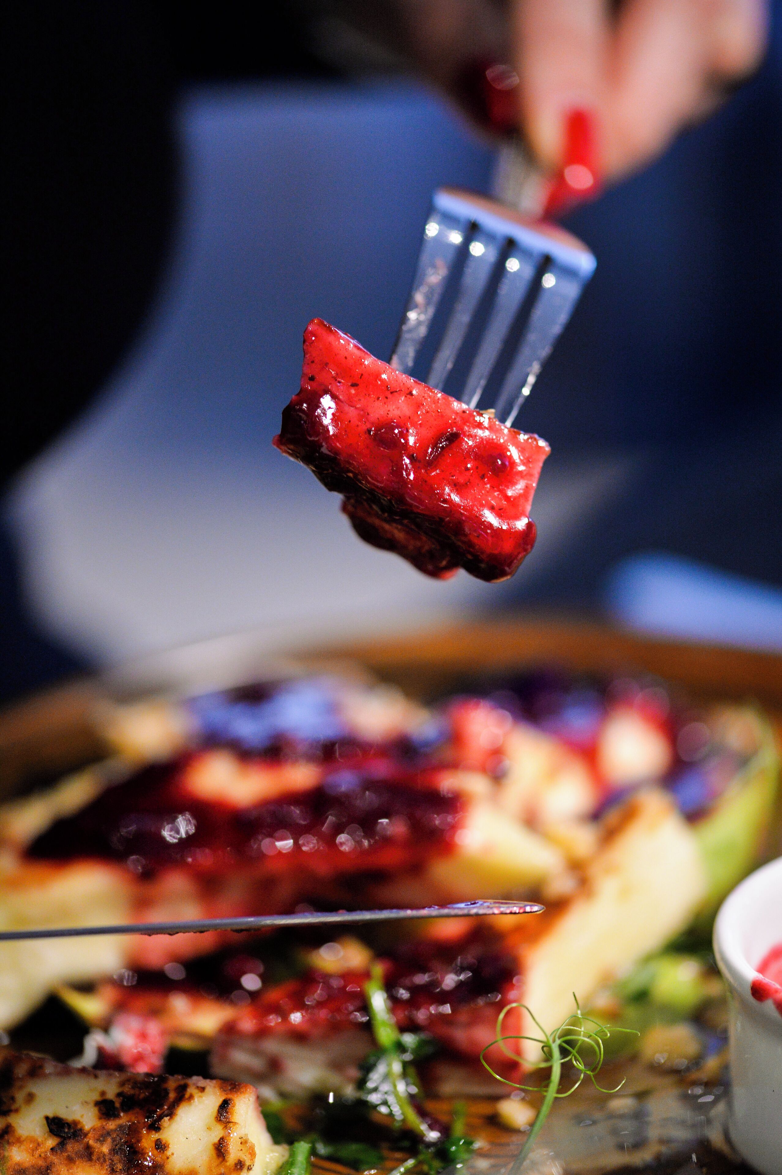 A close-up of a fork holding a piece of food with a red sauce, possibly a dessert or appetizer, on a plate with other food items in the background.