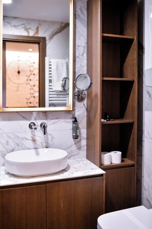 Un baño con lavamanos de mármol, armarios de madera, un espejo y una estantería con artículos de aseo.