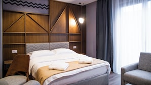 Una habitación de hotel con una cama grande, cabecera de madera con diseños geométricos, un rincón de descanso con dos sillones y un ventanal con cortinas.