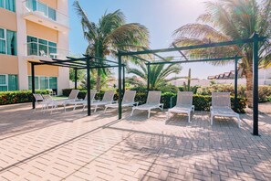 A covered outdoor seating area with white lounge chairs and a palm tree backdrop.