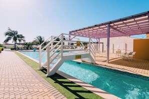 A swimming pool with a white diving platform, a covered seating area, and a brick walkway. - Spacious Condo with Pool Access in Noord (Noord)