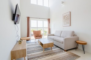 Un salon moderne comprenant un canapé, une table basse et un fauteuil, un téléviseur à écran plat fixé au mur et une grande fenêtre avec des rideaux.