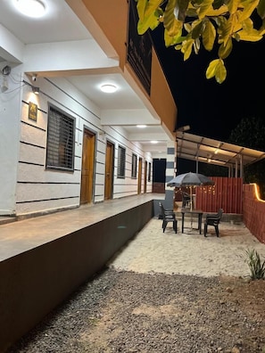 A covered outdoor seating area with tables and chairs, a wooden fence, and a gravel ground. - Crown hotel (Shrivardhan)