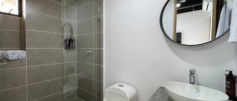 Une salle de bain moderne avec une douche à parois de verre, des toilettes blanches et un grand lavabo surmonté d’un grand miroir rond.