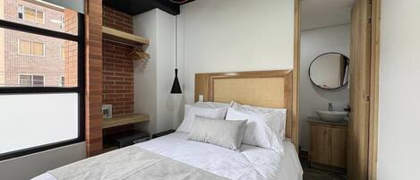 Un dormitorio con cama, pared de ladrillo, ventana, cabecera de madera, mesita de noche, lámpara, espejo y baño con lavamanos.