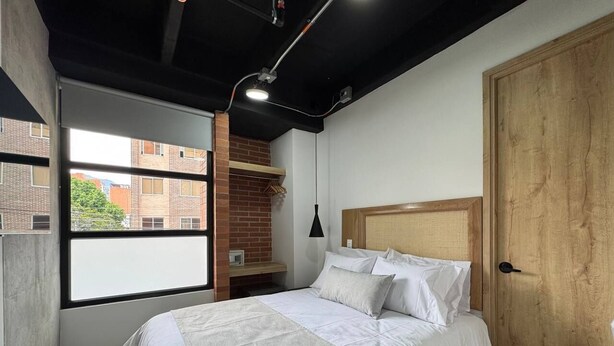 A bedroom with a bed, a window, a brick wall, a wooden door, and a hanging light fixture. - Akela Park (Medellín)