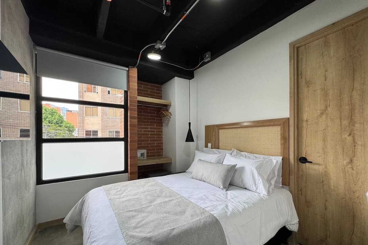 A bedroom with a bed, a window, a brick wall, a wooden door, and a hanging light fixture.