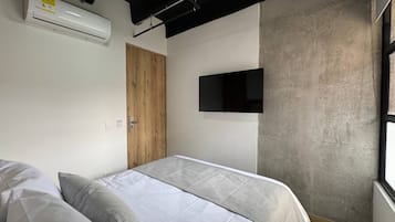A bedroom with a bed, pillows, an air conditioner, a wooden door, a TV mounted on the wall, and a window.