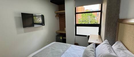 A modern hotel room with a bed, a flat-screen TV mounted on the wall, a small desk with a chair, and a window with a view of buildings and greenery.