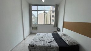 Una habitación pequeña y poco amoblada con una cama, una silla y una ventana con vistas a edificios.