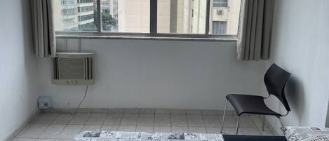 Une pièce avec une grande fenêtre, une chaise et un lit sur lequel est étalé un journal par terre.