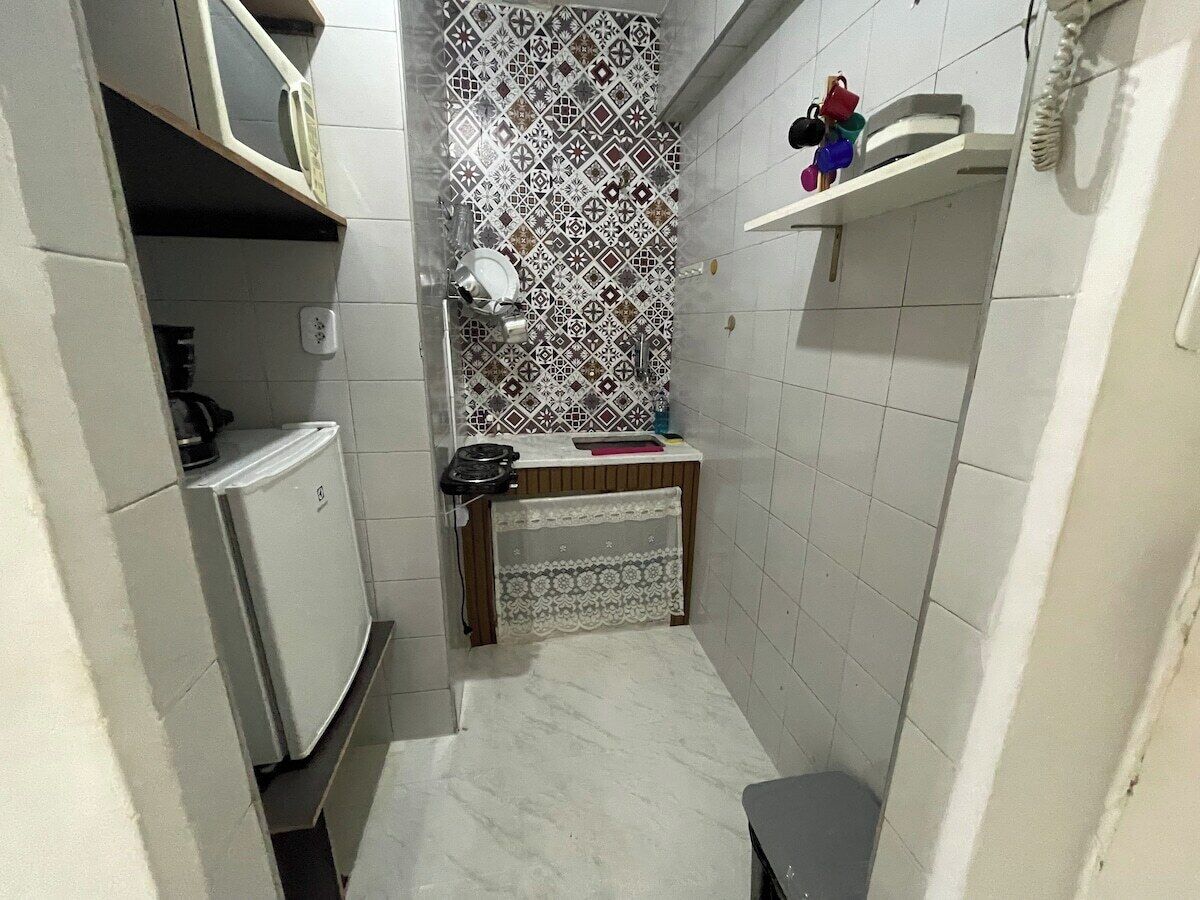 Un baño pequeño y estrecho con azulejos blancos, un salpicadero decorativo, un inodoro, un lavabo y un estante pequeño con artículos de aseo.