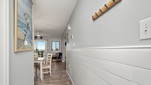 A hallway with a wooden floor, a dining area with a table and chairs, and a gray wall with a light switch and a wooden object. - Beach Retreat: Oceanfront Upper Level Corner Unit Close to Community Pool (Oak Island)
