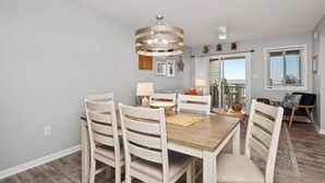 A dining area with a wooden table, white chairs, and a hanging light fixture. - Beach Retreat: Oceanfront Upper Level Corner Unit Close to Community Pool (Oak Island)