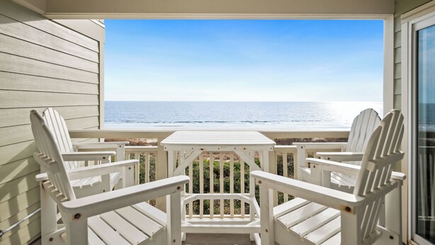 A balcony with white Adirondack chairs and a table, overlooking the ocean. - Beach Retreat: Oceanfront Upper Level Corner Unit Close to Community Pool (Oak Island)