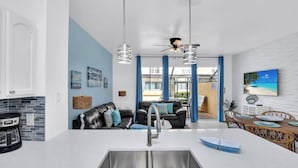 Een moderne keuken met een centraal eiland, een eethoek met een houten tafel en stoelen, en een woonkamer met een bank en blauwe gordijnen.