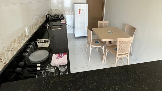 A compact kitchen with a black countertop, white tiled backsplash, a white refrigerator, and a dining area with a table and chairs. - Casa Corais Beach Francês (Marechal Deodoro)