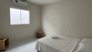 A minimalist bedroom with a bed, a small wooden nightstand, a window, and an air conditioning unit. - Casa Corais Beach Francês (Marechal Deodoro)