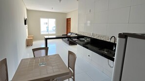 A modern kitchen with white tiles, a dining area with a table and chairs, and a view of the outside through a window. - Casa Corais Beach Francês (Marechal Deodoro)