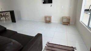 A modern living room with a dark sofa, a flat-screen TV mounted on the wall, and two small wooden side tables. - Casa Corais Beach Francês (Marechal Deodoro)