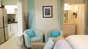 Ein Schlafzimmer mit zwei hellblauen Sesseln, einem kleinen weißen Tisch, einem Bett mit weißer und violetter Bettwäsche, einer weißen Kommode und einem gerahmten Bild an der Wand.