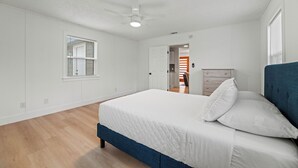 Ein Schlafzimmer mit einem Bett, einem Deckenventilator, einem Fenster mit Jalousien und einer Tür zu einem weiteren Raum.