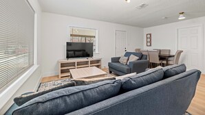 Ein modernes Wohnzimmer mit grauem Sofa, einem Flachbildfernseher auf einem Holzboden, einem Esstisch mit Stühlen und einer Kristallleuchter.