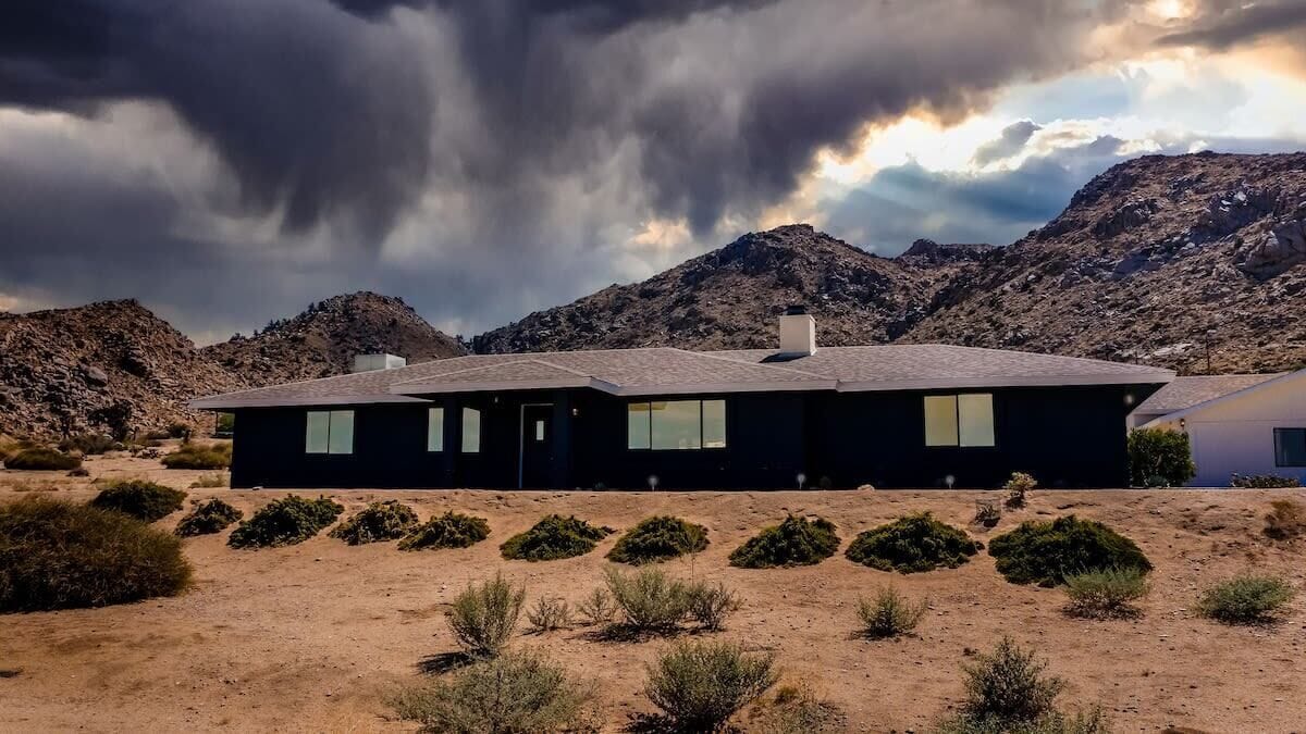A modern two-story house with a flat roof, surrounded by desert vegetation and mountains in the background.