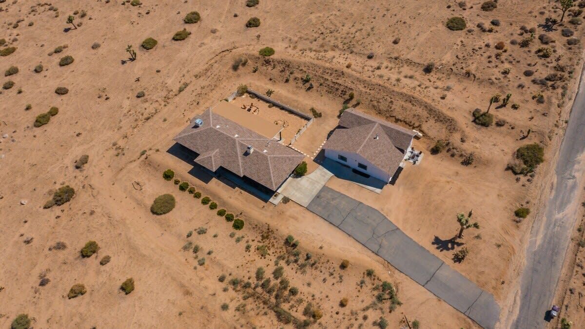 An aerial view of a residential area with two houses, a paved driveway, and sparse vegetation.