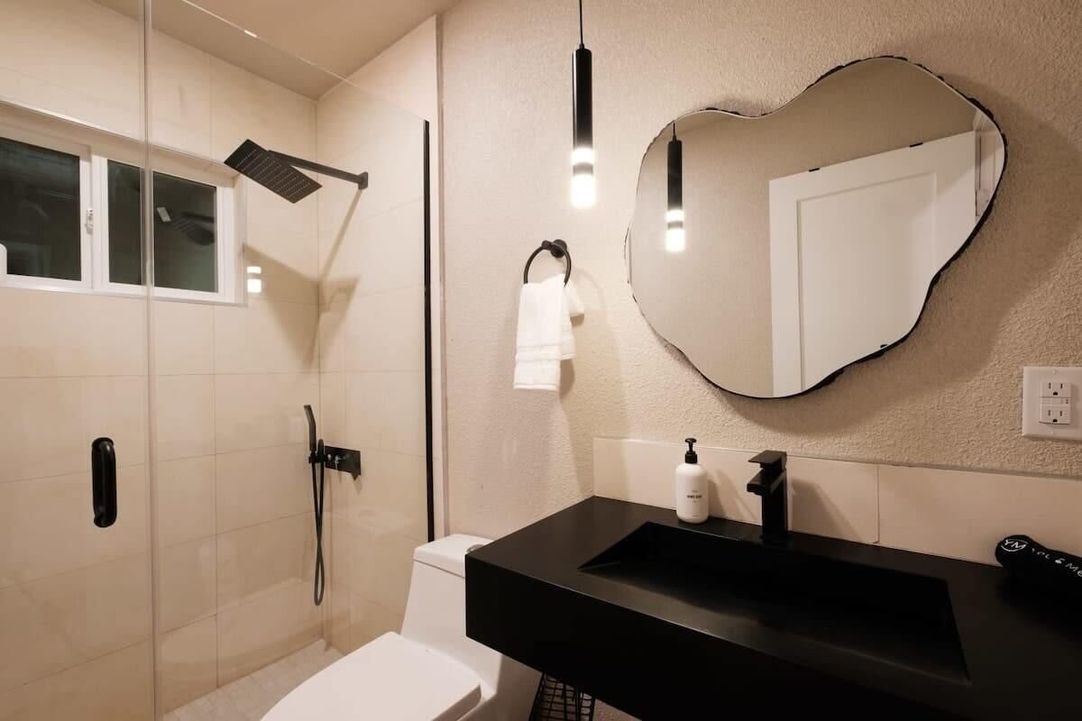 Un baño moderno con ducha de vidrio, lavamanos negro y espejo con un original marco con forma de corazón.