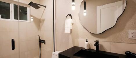 Un baño moderno con ducha de vidrio, lavamanos negro y espejo con un original marco con forma de corazón.