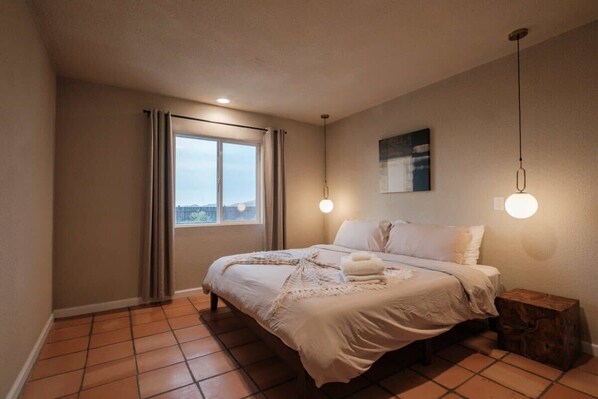 A bedroom with a large bed, a window with curtains, a wooden nightstand, and a wall-mounted light fixture.