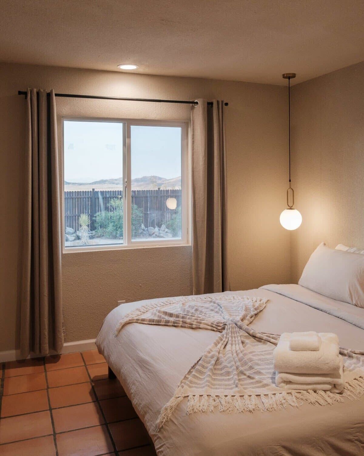 Habitación de hotel con cama, ventana con cortinas, lámpara colgante y vistas al exterior.