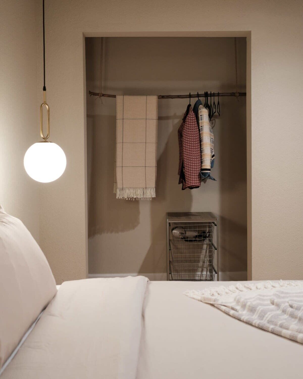 A bedroom with a bed, a hanging light fixture, a closet with clothes and a towel, and a metal rack with items.