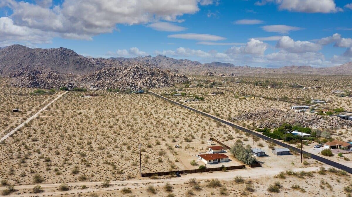 A desert landscape with sparse vegetation, a few buildings, and mountains in the background.
