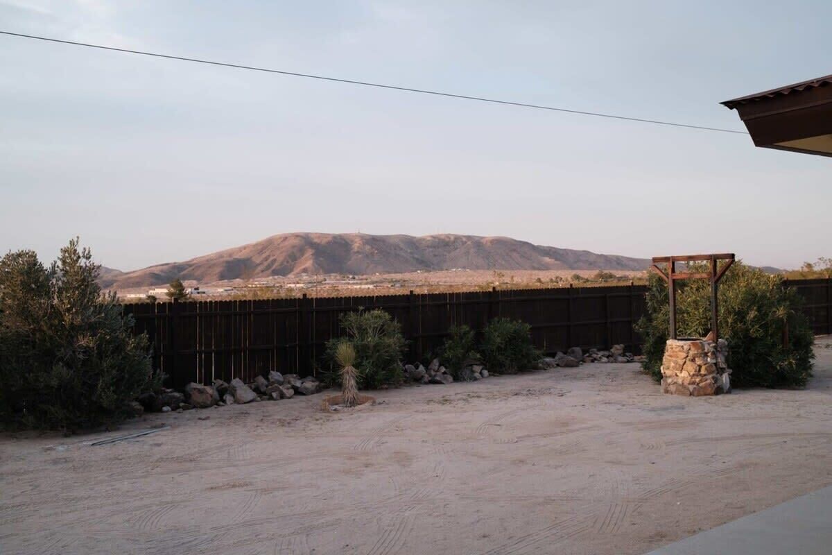 A desert landscape with a mountain in the background, a fenced area with sparse vegetation, and a stone structure.