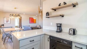 Une cuisine moderne avec des meubles blancs, un lave-vaisselle, une machine à café et un grille-pain. Deux étagères en bois sont fixées au mur, au-dessus du plan de travail.