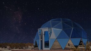 Une structure en forme de dôme géodésique dotée d’une grande fenêtre offrant une vue dégagée, installée sur une plateforme avec un paysage désertique en arrière-plan.