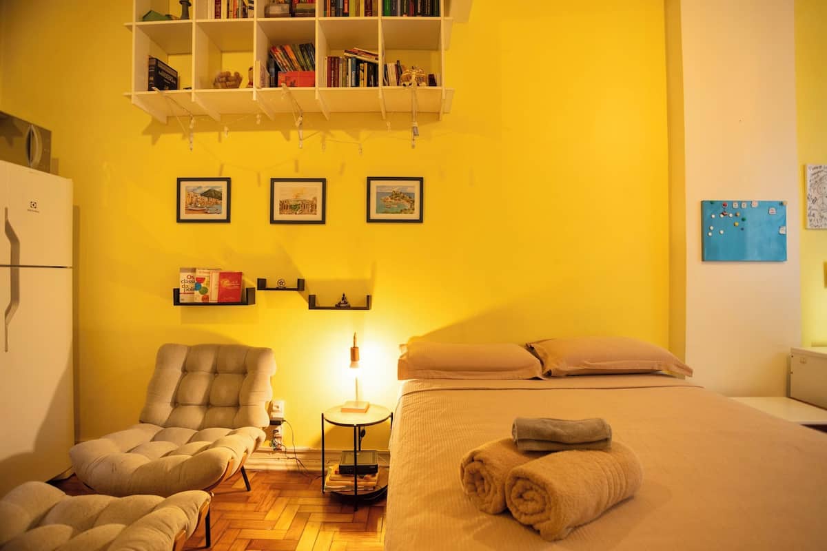 A room with a bed, two armchairs, a bookshelf, and framed pictures on the wall.