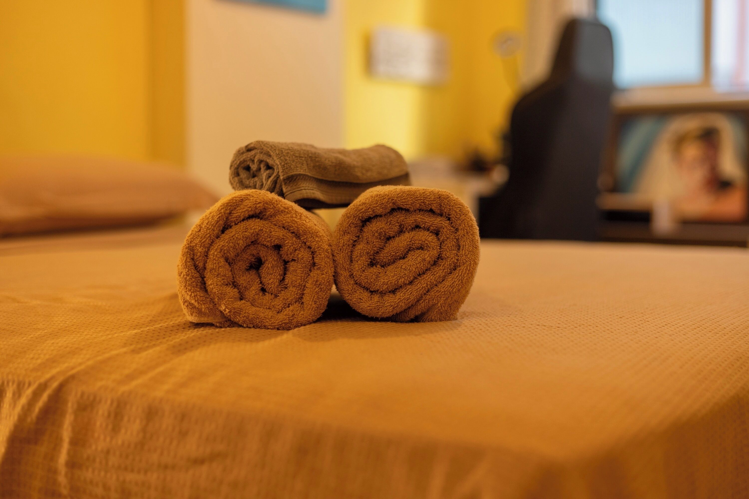 A bed with two rolled towels and a chair in a room with yellow walls.
