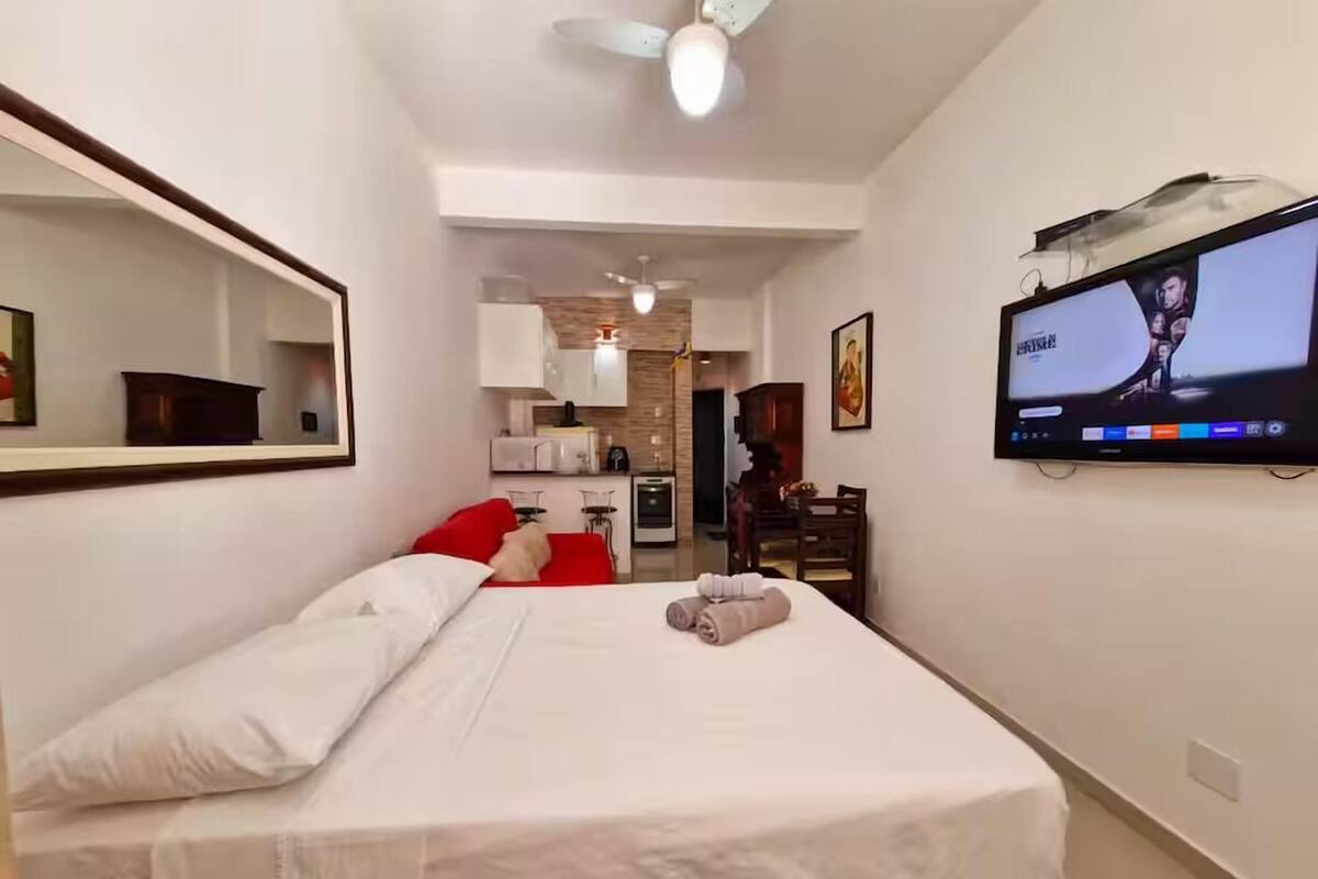 A hotel room with a bed, a TV mounted on the wall, a ceiling fan, and a kitchen area in the background.