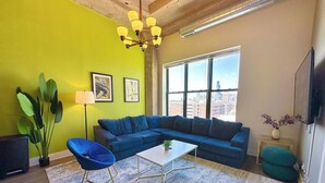 Una sala de estar con un sofá modular, una mesa de centro, un sillón azul, una lámpara de pie, una planta grande en maceta y vista a la ciudad a través de la ventana.