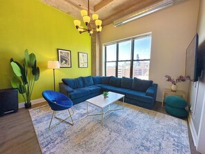 A living room with a sectional sofa, a coffee table, a blue chair, a floor lamp, a large potted plant, and a city view through the window.